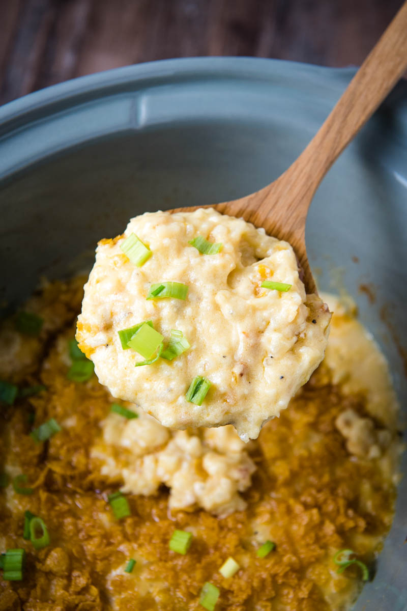 serving of slow cooker funeral potatoes, sprinkled with green onions, on wooden spoon over CrockPot