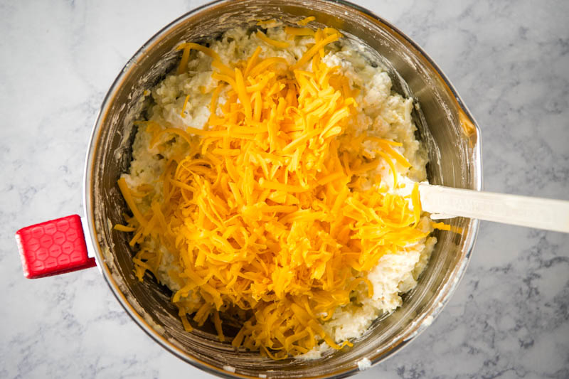 stirring cheddar cheese into hash brown mixture in metal mixing bowl with spatula