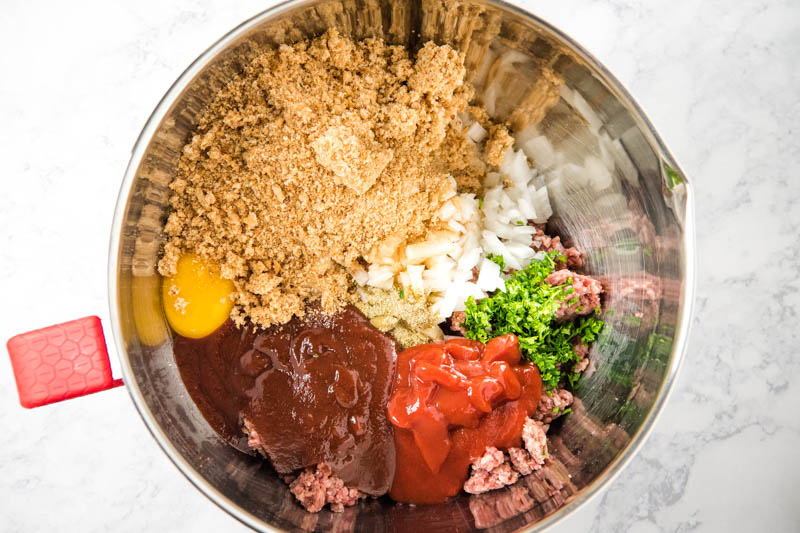 traditional meatloaf ingredients, including ground beef, eggs, onion, garlic, parsley, seasoned salt, ketchup, bbq sauce, and bread crumbs in metal mixing bowl