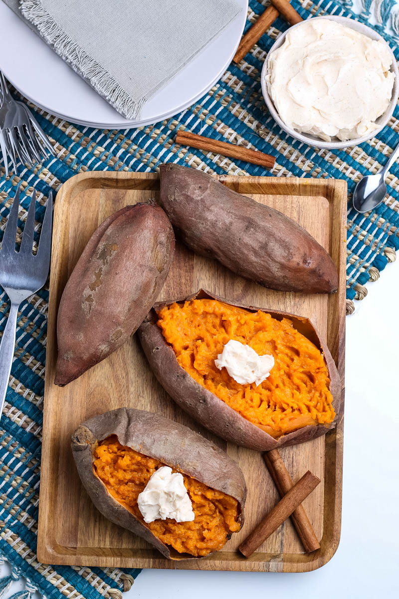pressure cooked sweet potatoes on wooden cutting board with cinnamon sticks, on blue woven placemat with white plates, fork, spoon, and small bowl of whipped cinnamon butter