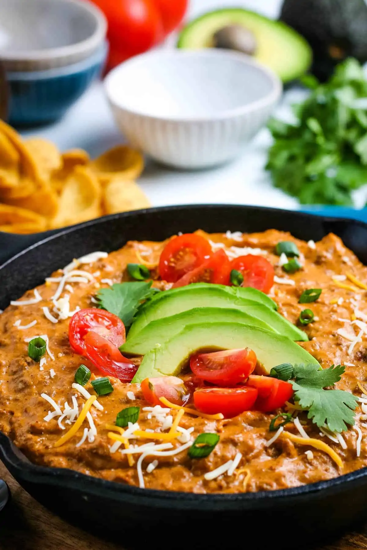 close up of chili dip, topped with sliced avocado, cilantro, and fresh diced tomatoes, in cast iron skillet