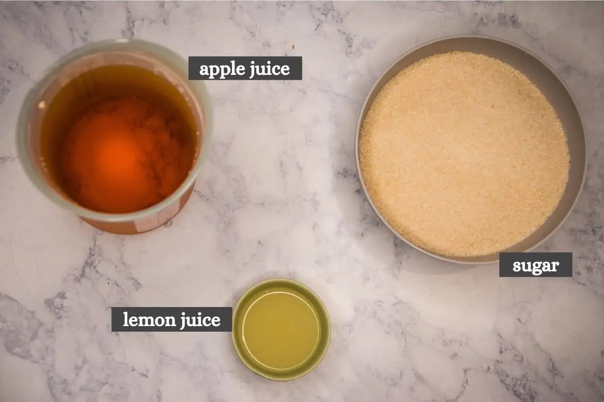 3 ingredients for apple jelly recipe, including apple juice, lemon juice, and sugar on white marble countertop
