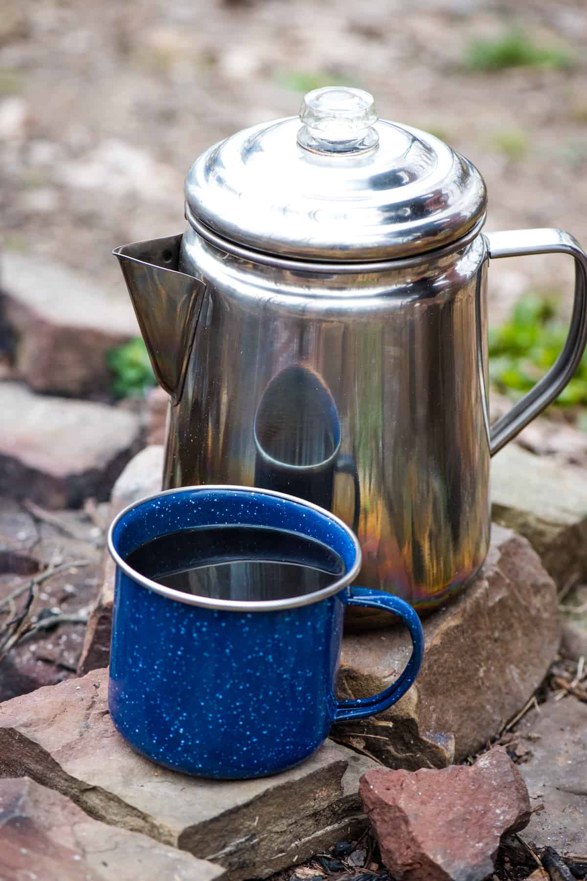 blue enamelware mug full of campfire coffee with stainless steel percolator on campfire ring rocks