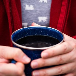 person with red jacket holding a blue enamelware mug of freshly brewed campfire coffee