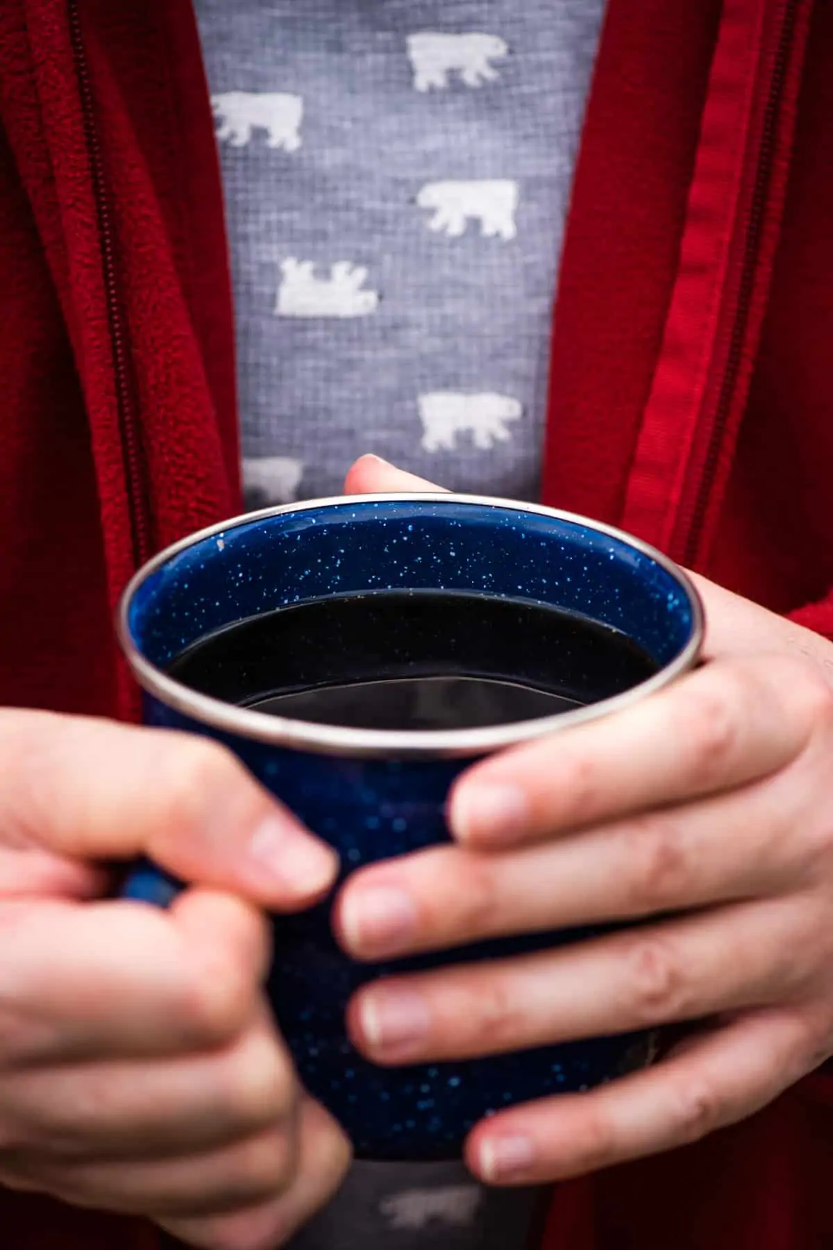 person with red jacket holding a blue enamelware mug of freshly brewed campfire coffee