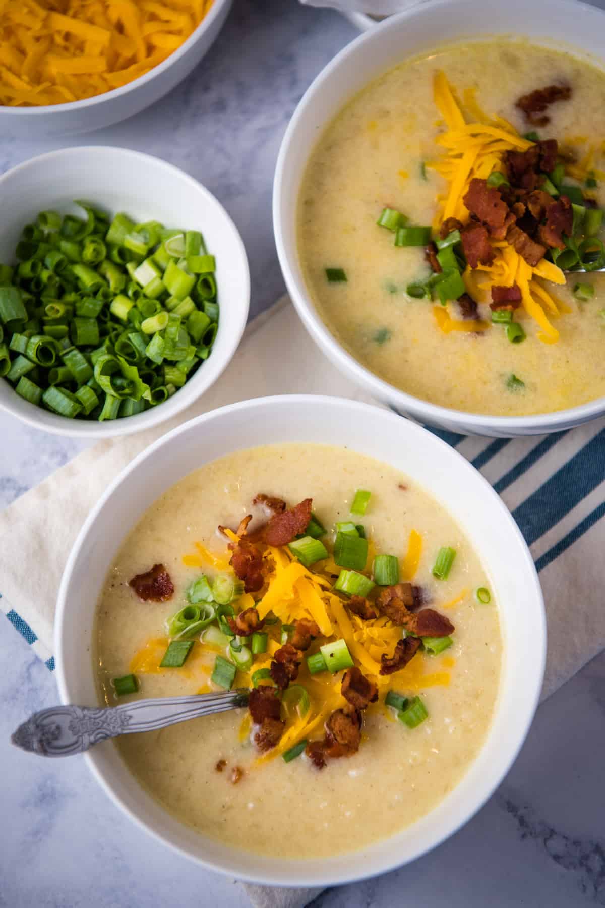 white bowls of Instant Pot potato soup topped with crumbled bacon, shredded cheese, and green onions, on white marble countertop with white blue striped kitchen towel