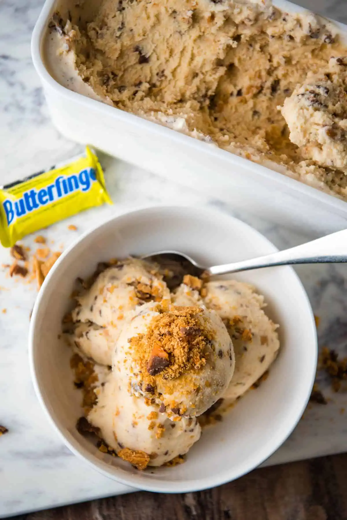 homemade Butterfinger ice cream topped with crushed Butterfinger bars in white bowl with spoon next to ice cream container