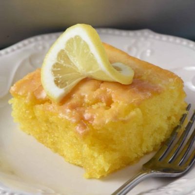 sliced square of easy lemon poke cake with lemon glaze and topped with a fresh lemon slice on a small white saucer