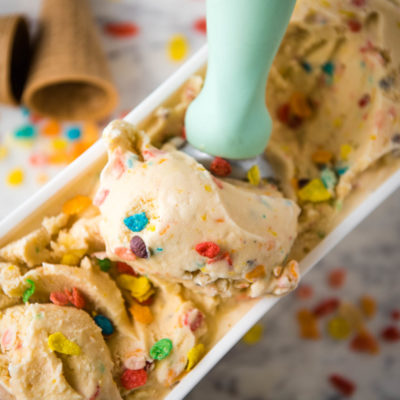 mint green scoop in white ice cream container filled with Fruity Pebbles ice cream on white marble cutting board with sugar cones and Fruity Pebbles cereal