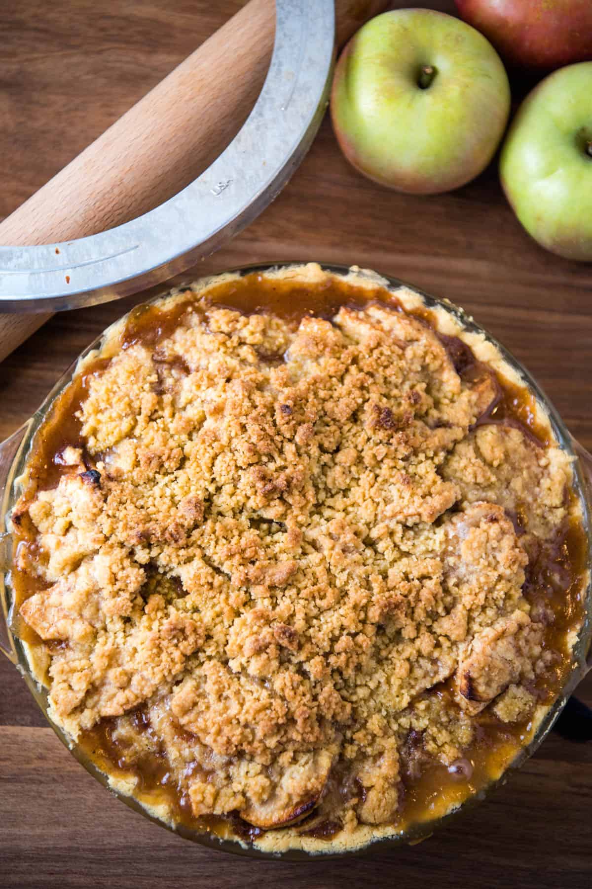 apple pie in glass pie plate with perfectly cooked edges on wooden countertop with pie shield and rolling pin