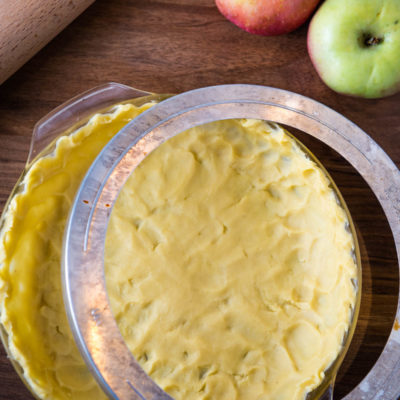 unbaked pie crust with metal pie shield on wooden countertop with apples and rolling pin