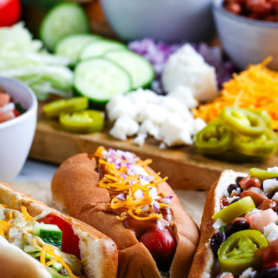 loaded slow cooker hot dogs next to all of the toppings lined out on a large wooden cutting board
