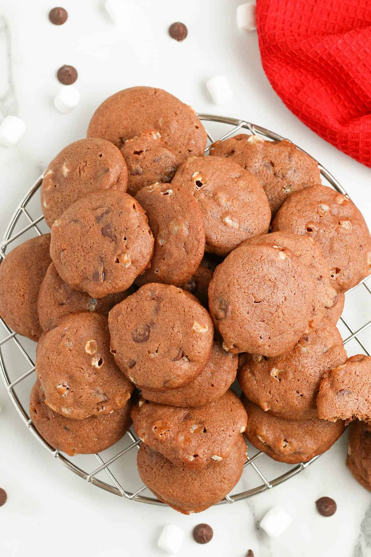 hot chocolate cookies piled up on a silver wire cooling rack on a white countertop with chocolate chips and marshmallows