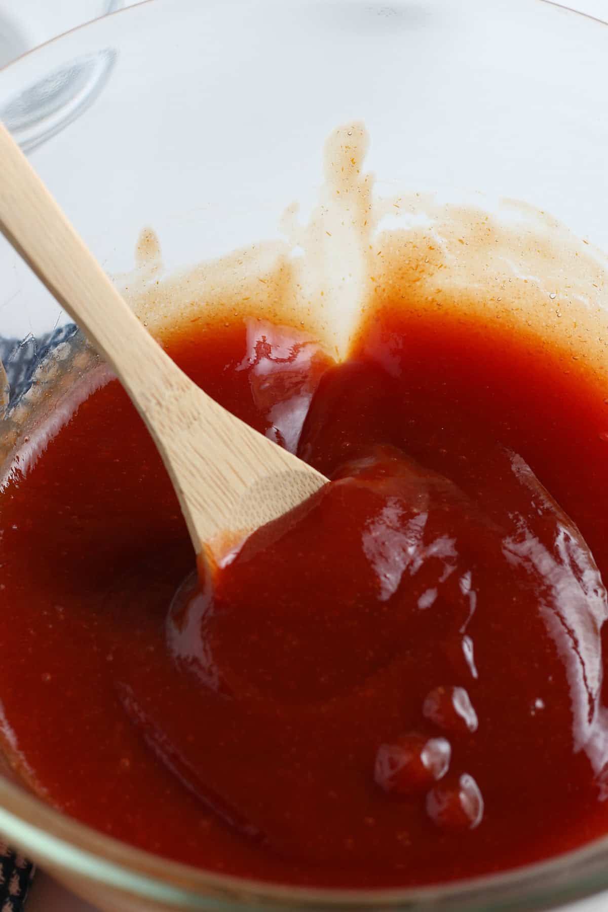 closeup photo showing a large wooden spoonful of homemade barbecue sauce in a large glass bowl full of homemade bbq sauce