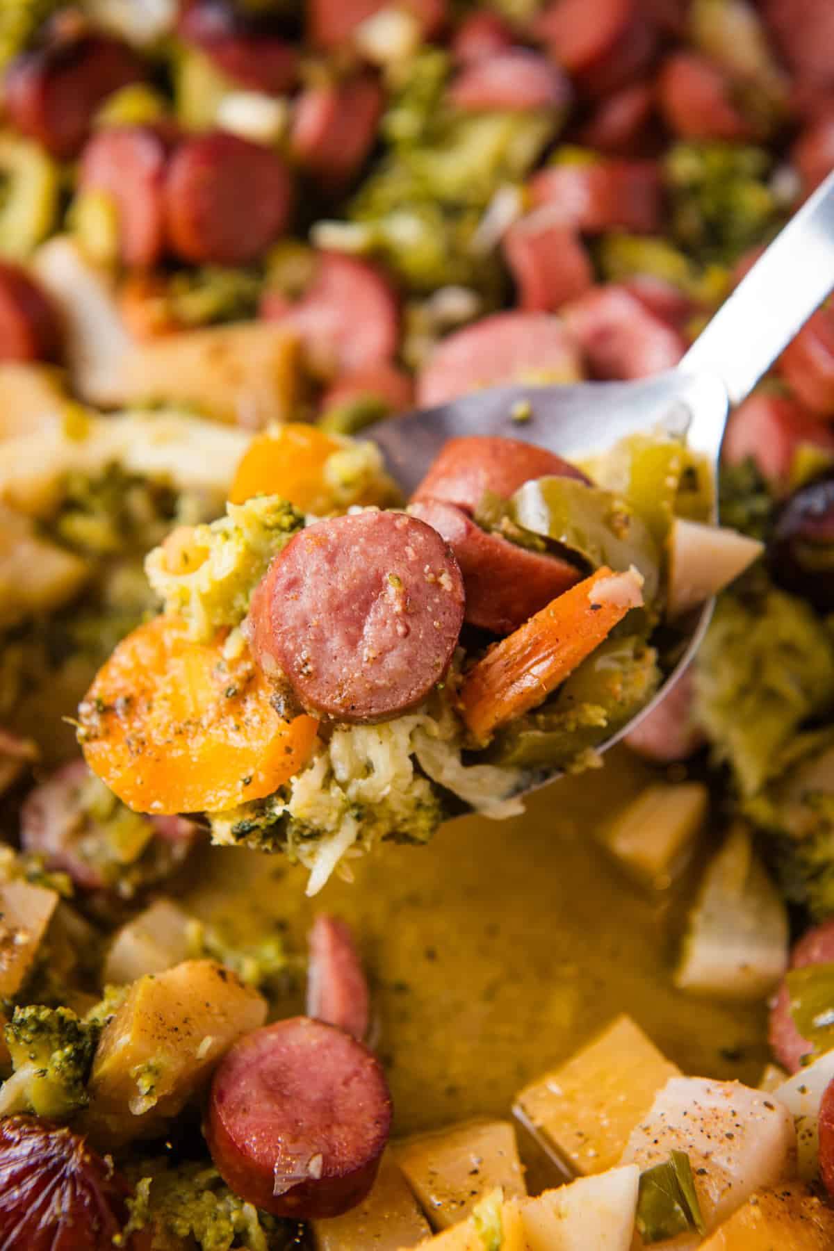 closeup photo showing a heaping spoonful of sausage and vegetables over a large baking pan full of sheet pan sausage and veggies