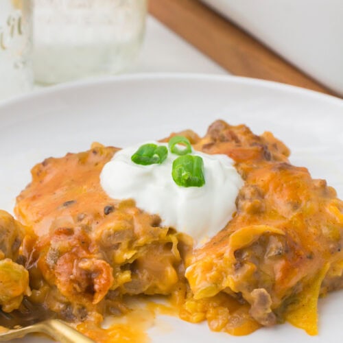 beef enchilada casserole on a small white plate with sour cream and green onions on top for garnish