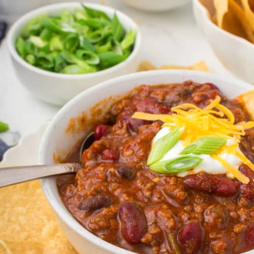 chili con carne in a small white bowl with a dollop of sour cream, chopped green onions and shredded cheese on top