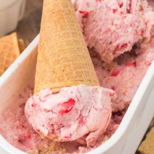 strawberry cheesecake ice cream in a waffle cone tipped upside down inside of a tub of strawberry cheesecake ice cream