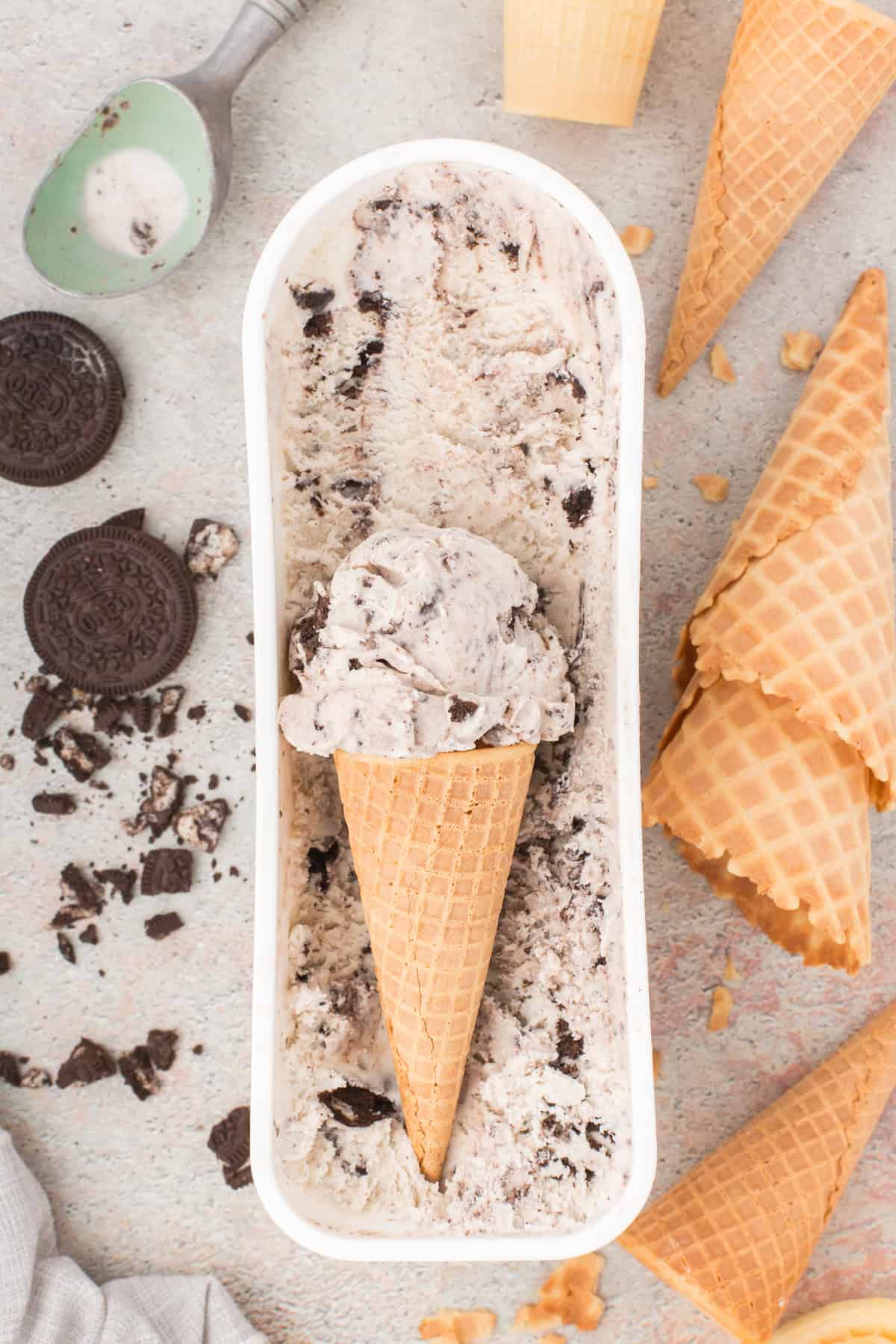 overhead shot showing best homemade Oreo ice cream in a large rectangular ice cream tub and a scoop inside of a waffle cone laying on top