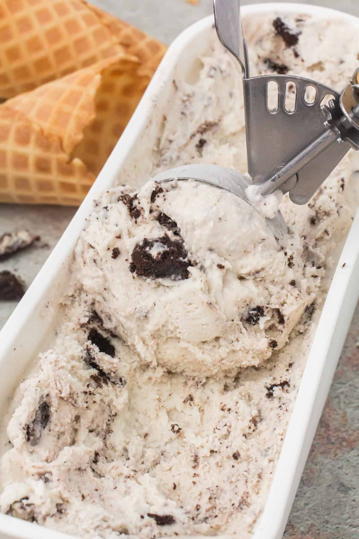closeup shot showing an ice cream scooper scraping homemade cookies and cream ice cream out of a large white rectangular ice cream container