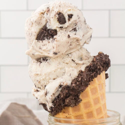 closeup shot showing two scoops of homemade cookies and cream ice cream in a waffle cone with crumbled up Oreo cookie pressed around the rim of the cone