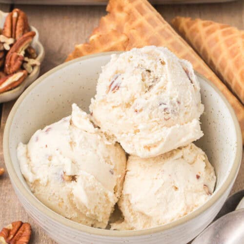 three scoops of homemade butter pecan ice cream in a small gray decorative bowl with waffle cones in the background