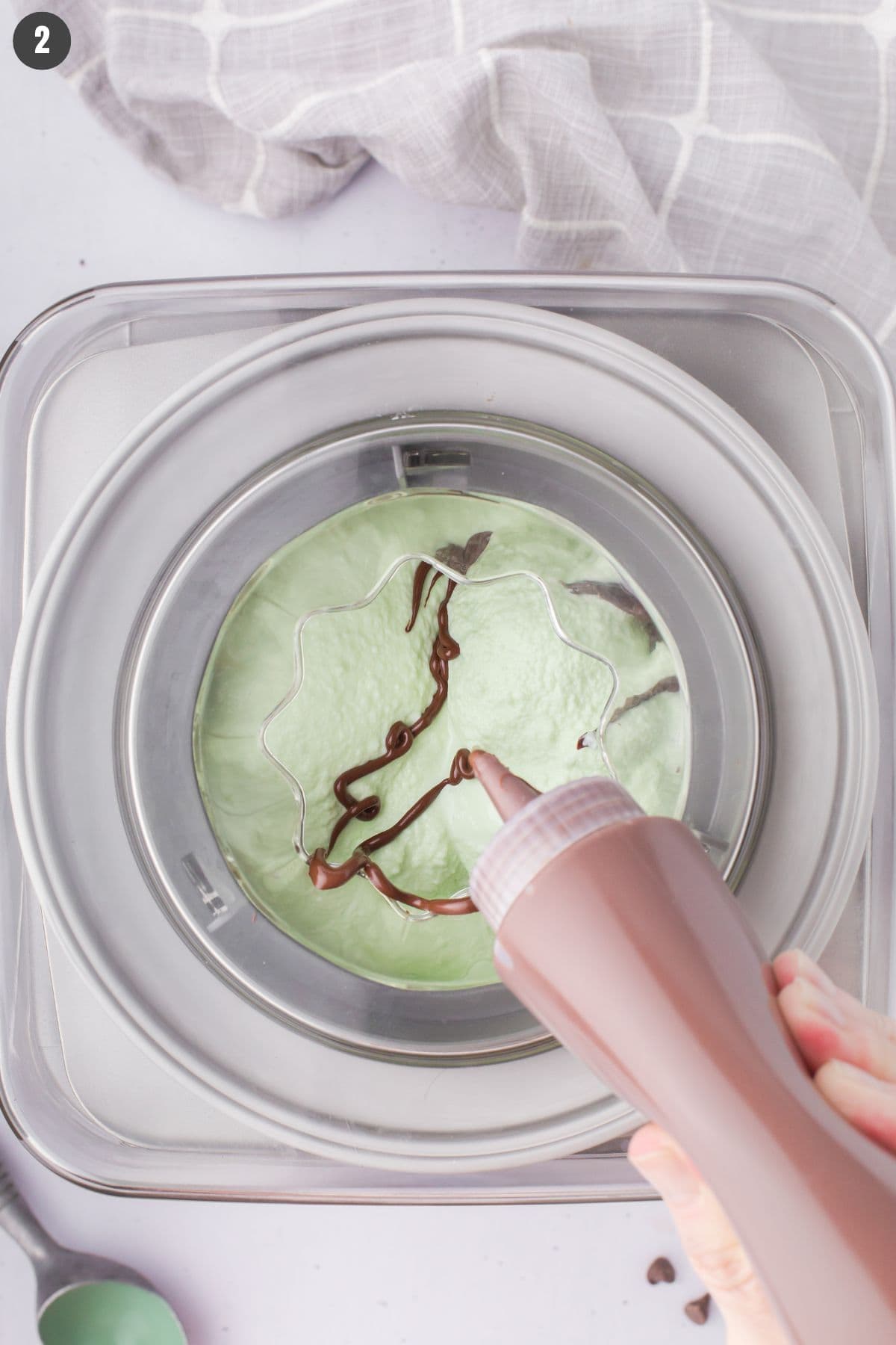 closeup shot showing melted chocolate being squeezed out of a plastic bottle and into the homemade mint chocolate chip ice cream in a churn-style ice cream maker