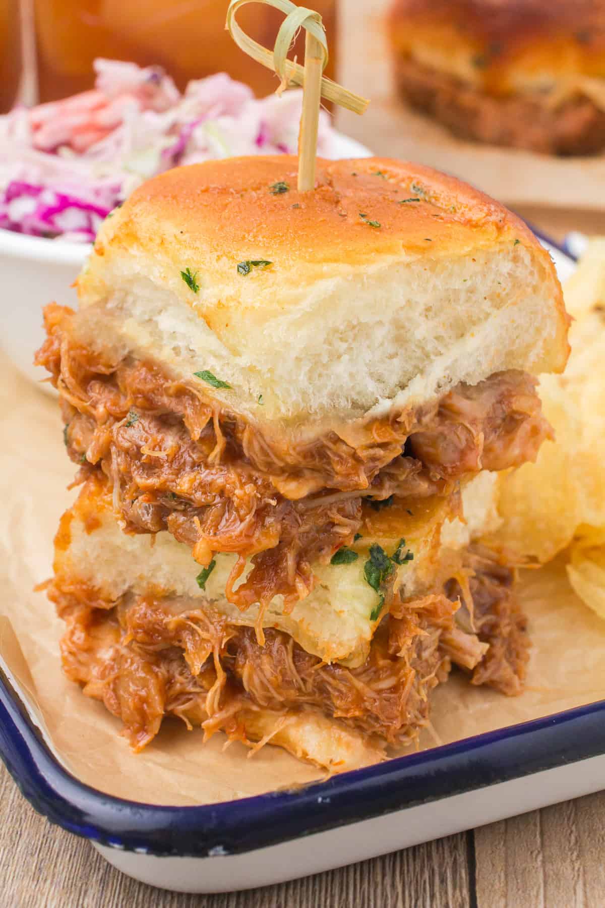 stacked slow cooker bbq chicken sliders on Hawaiian rolls on enamel baking sheet with potato chips and cole slaw