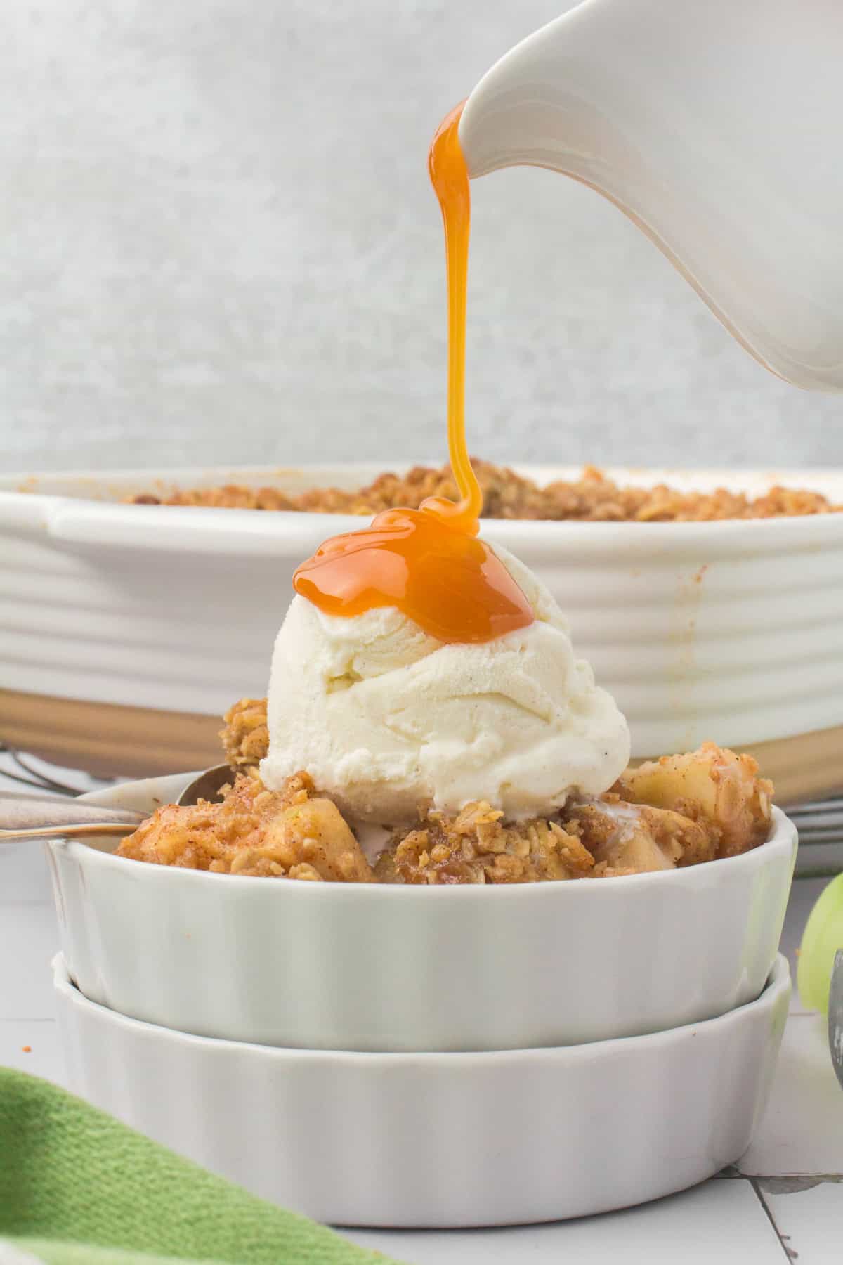 closeup shot showing homemade apple crisp in a small white ramekin dish with a scoop of vanilla ice cream on top and caramel sauce being drizzled out of a white cream pitcher