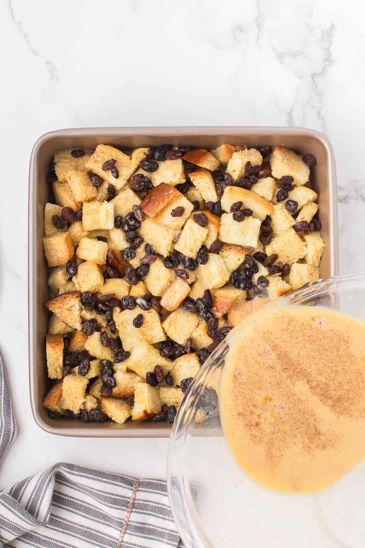 overhead shot showing a glass measuring cup full of egg custard being poured all over the top of bread pudding in a 9x9 pan