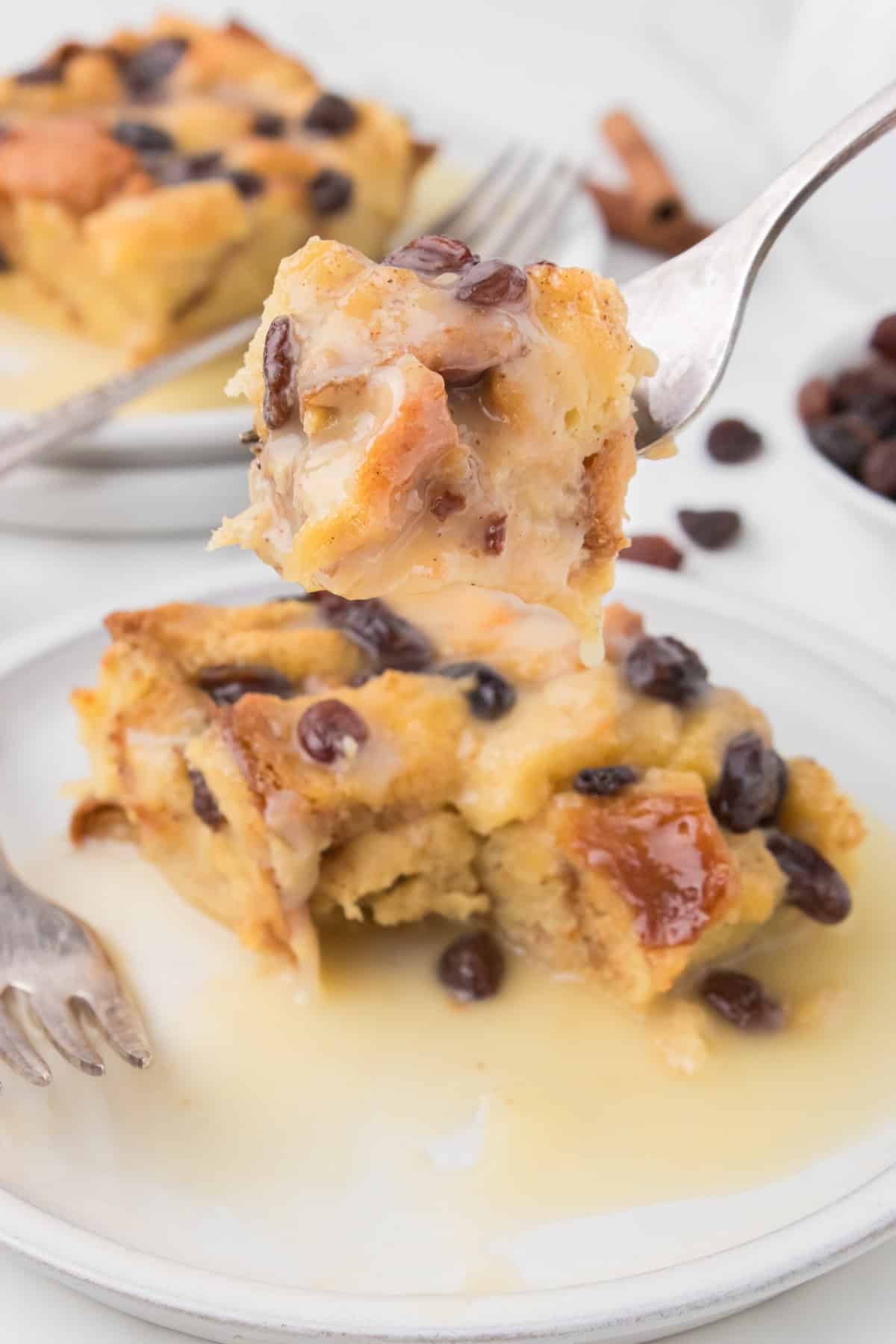 closeup shot showing a bite of bread pudding on a fork over top of a sliced square of bread pudding with vanilla sauce on a small white decorative plate