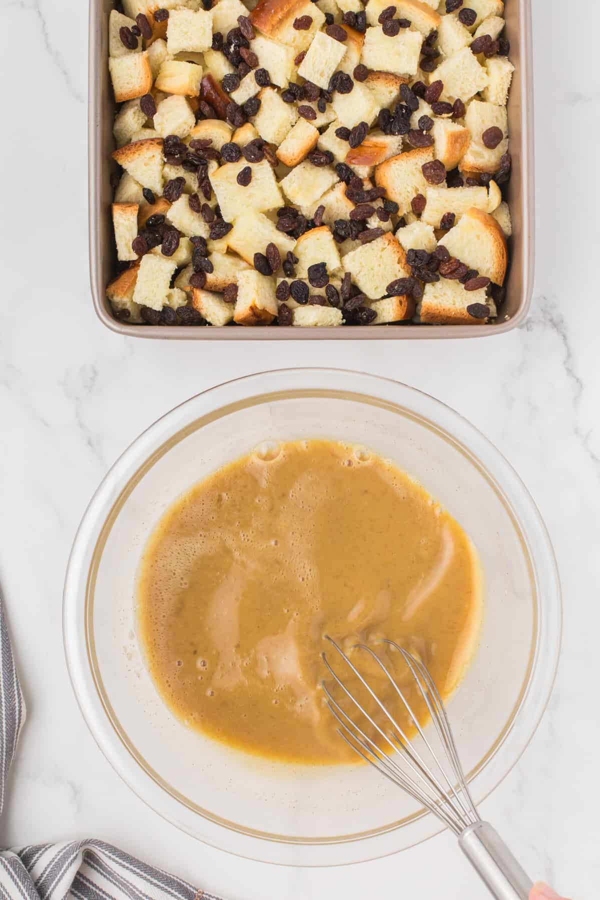 whisking the egg mixture in a small glass mixing bowl with a 9x9 pan full of homemade bread pudding on a white countertop