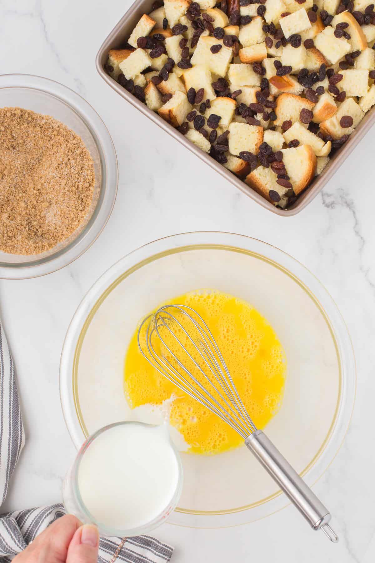 pouring cream into the egg custard in a small glass mixing bowl with a 9x9 pan full of homemade bread pudding dessert in the background